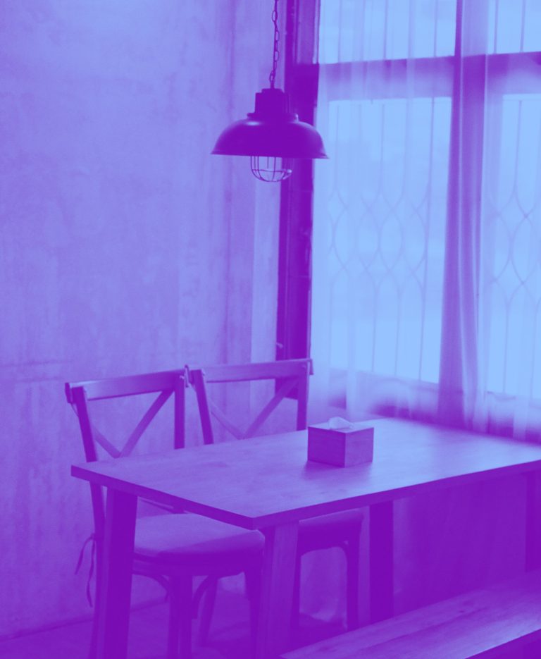 Un interno spoglio con un tavolo di legno, due sedie e una lampada sospesa, in duotone viola e azzurro